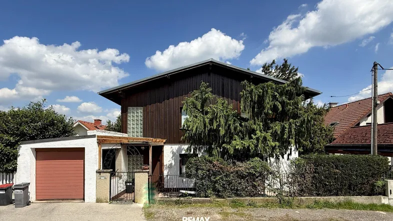 Traditionelles Einfamilienhaus mit Holzfassade und integrierter Garage unter blauem Himmel.