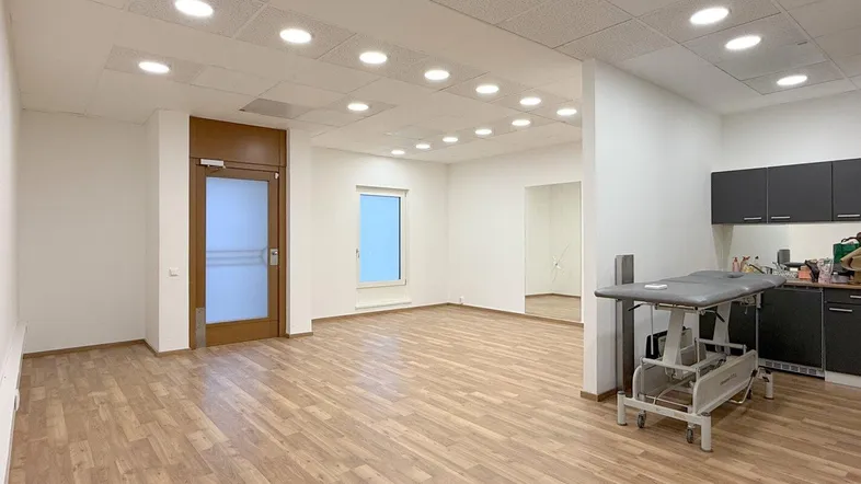 Geräumiger Büroraum mit hellem Laminatboden, Einbaudeckenleuchten und einer Behandlungsliege im Hintergrund.