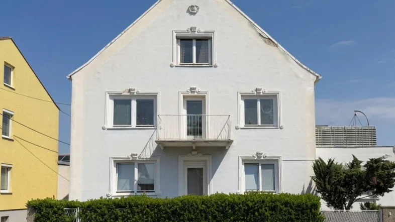 Klassisches Wohnhaus mit weißer Fassade, kleinem Balkon und gepflegter Hecke.