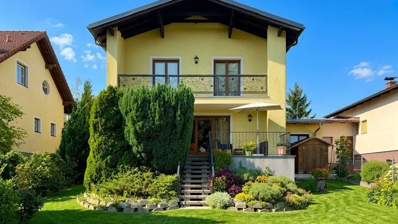Gelbes Einfamilienhaus mit gepflegtem Garten, Terrasse und Balkon unter blauem Himmel.