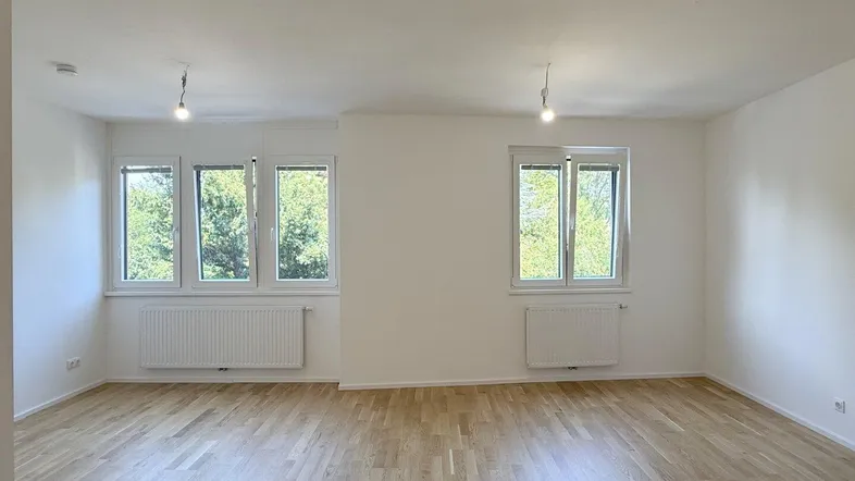 Helles Zimmer mit Holzboden und Blick ins Grüne, ausgestattet mit Heizkörpern unter den Fenstern.