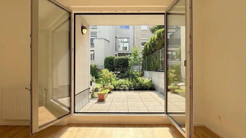 Helles Zimmer mit Holzboden und direktem Zugang zu einer gepflasterten Terrasse und einem kleinen Garten.
