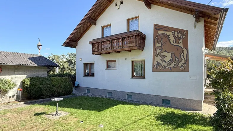 Ansicht des Hauses mit traditionellem Balkon und einem gepflegten Vorgarten unter blauem Himmel.
