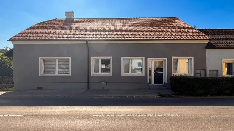 Graue Fassade eines Einfamilienhauses mit roten Dachziegeln unter blauem Himmel.