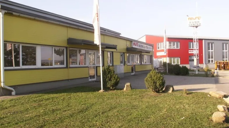 Gebäude mit gelber Fassade und rotem Anbau, umgeben von einer Grünfläche und einem Parkplatz.