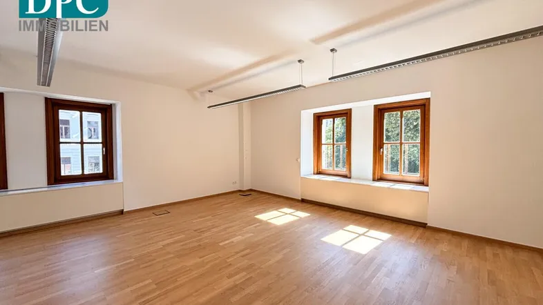 Geräumiger, leerer Büroraum mit hellem Parkettboden und mehreren Fenstern, die viel Tageslicht hereinlassen.