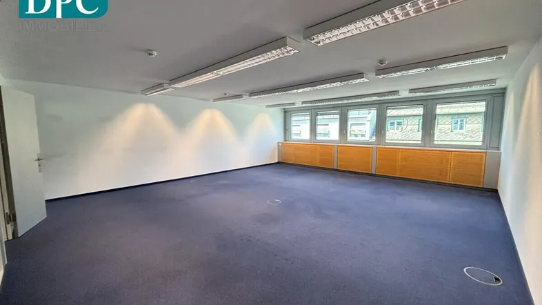 Geräumiges Büro mit blauem Teppichboden, großen Fenstern und Deckenbeleuchtung, bereit zur Nutzung.