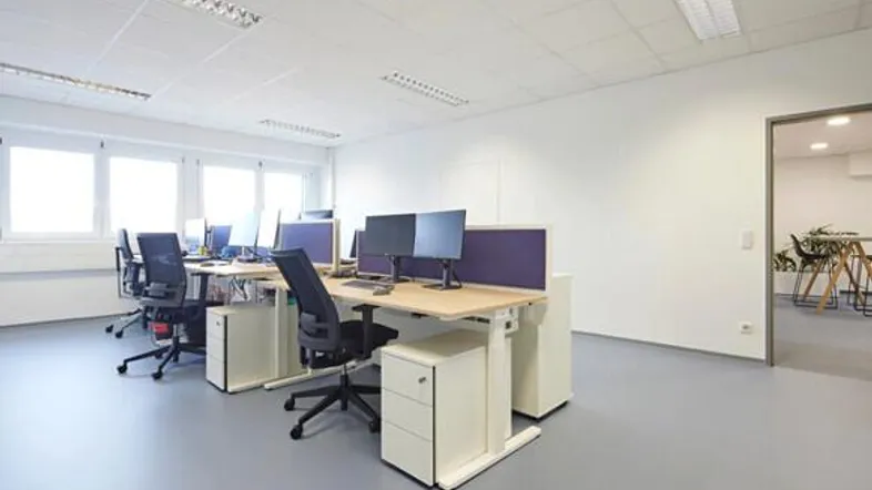 Helles und modernes Büro mit mehreren Arbeitsplätzen, großen Fenstern und höhenverstellbaren Schreibtischen.