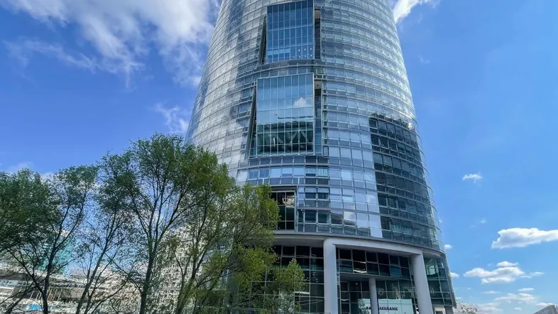 Beeindruckendes modernes Hochhaus mit Glasfassade und blauer Himmel im Hintergrund, umgeben von Bäumen.