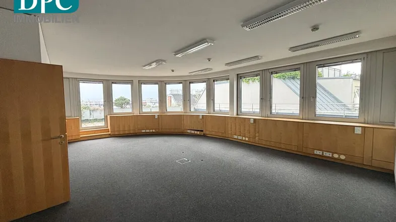 Großer, runder Büroraum mit vielen Fenstern, Teppichboden und Holzvertäfelung, ideal für offene Arbeitsbereiche.