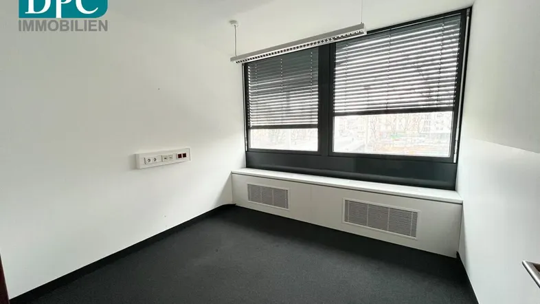 Heller Büroraum mit großen Fenstern, dunklem Teppichboden und weißen Wänden, ideal für konzentriertes Arbeiten.