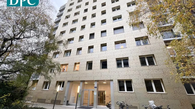 Mehrstöckiges Wohngebäude mit moderner Fassade, umgeben von Bäumen und Fahrradständern am Eingangsbereich.