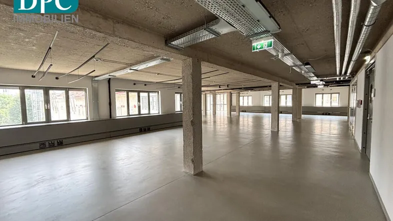 Großer, offener Büroraum mit Betonboden, sichtbaren Deckeninstallationen und vielen Fenstern für natürliches Licht.