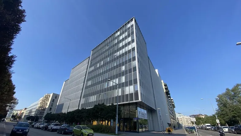 Modernes Bürogebäude mit Glasfassade und klarem Himmel, umgeben von einer städtischen Straße mit parkenden Autos.