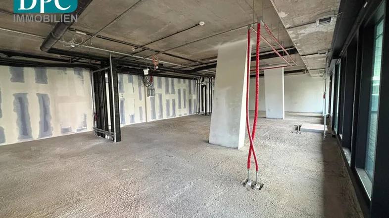 Großer, unfertiger Büroraum mit Betonboden und -decke sowie großen Fenstern, bereit für den Ausbau.