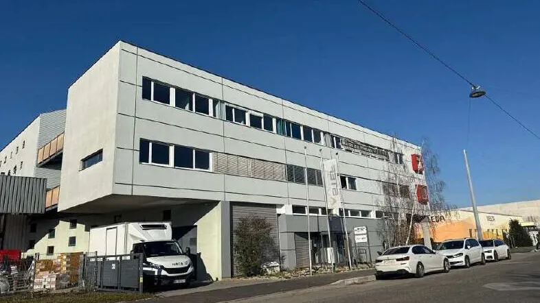 Modernes Bürogebäude mit mehreren Etagen, großen Fenstern und Parkplätzen entlang der Straße.