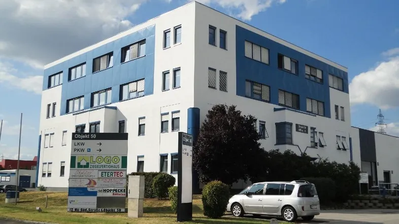 Modernes Bürogebäude mit weiß-blauer Fassade und Beschilderung für die Zufahrt unter bewölktem Himmel.