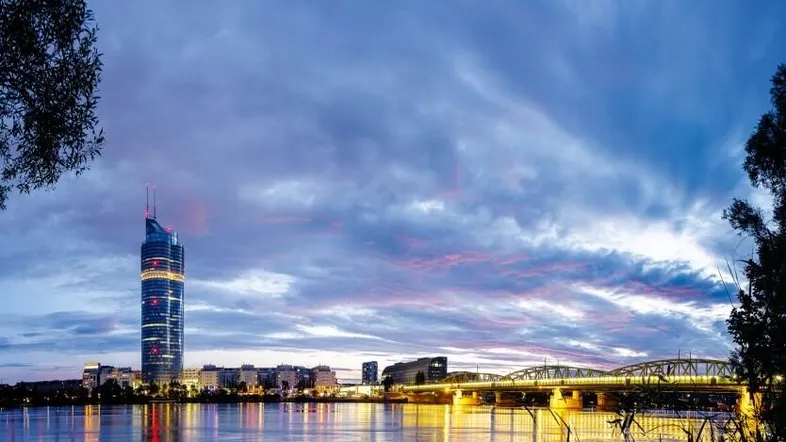 Abendliche Ansicht der Stadt mit beleuchtetem Wolkenkratzer, Fluss und Brücke unter dramatischem Himmel.