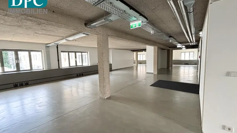 Großer, offener Büroraum mit Sichtbetondecke, Industriedesign und vielen Fenstern für natürliches Licht.