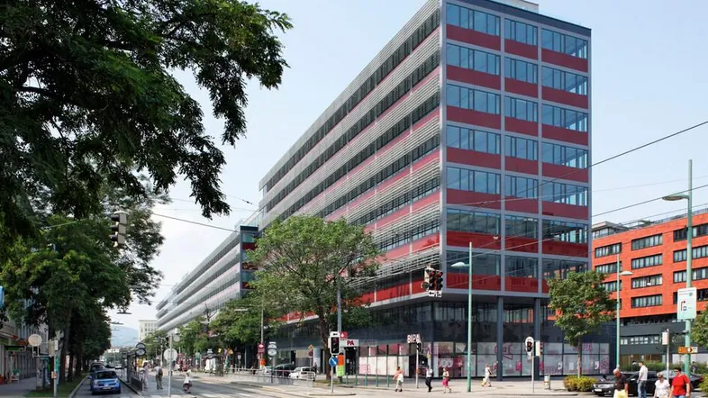 Modernes Bürogebäude mit vielen Fenstern an einer belebten Kreuzung mit Straßenbahnschienen und Bäumen.