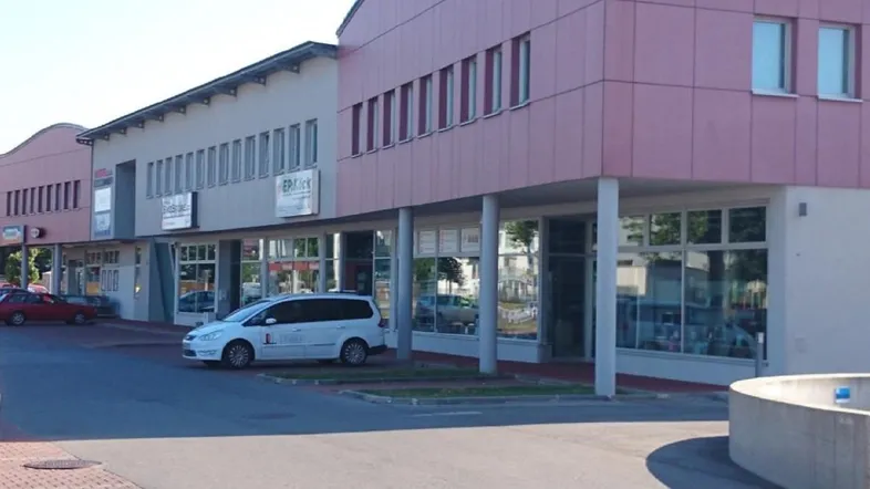 Gebäudeansicht mit mehreren Geschäften und Büros, einer modernen Fassade und Parkplätzen davor.