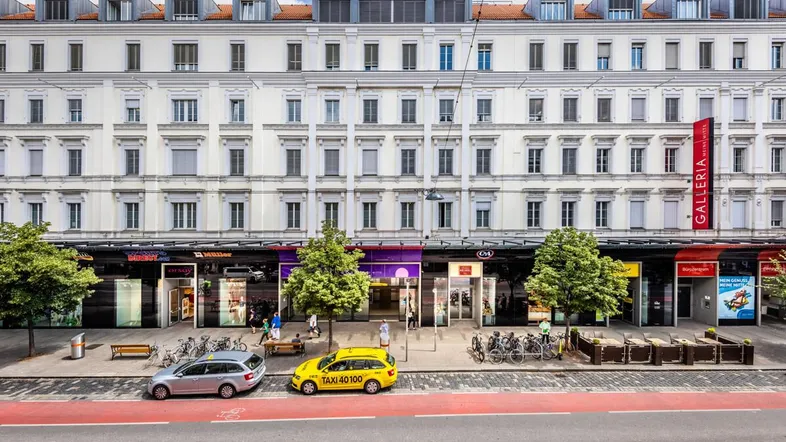 Ansicht eines klassischen Gebäudes mit Geschäften im Erdgeschoss und Wohnungen darüber, belebt durch Passanten und Fahrzeuge.