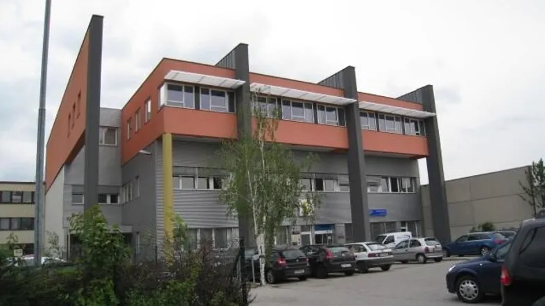 Modernes Bürogebäude mit markanter orange-grauer Fassade und Parkplätzen davor unter bewölktem Himmel.