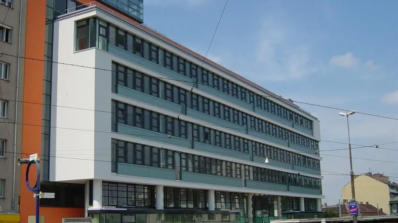 Modernes Bürogebäude mit mehreren Etagen, großen Fenstern und einer markanten orangefarbenen Seitenfassade in städtischer Umgebung.