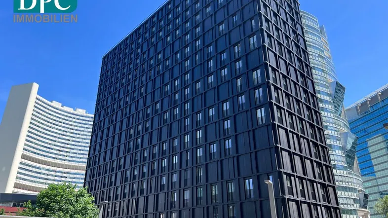 Modernes Hochhaus mit dunkler Fassade und vielen Fenstern unter blauem Himmel in einer städtischen Umgebung.