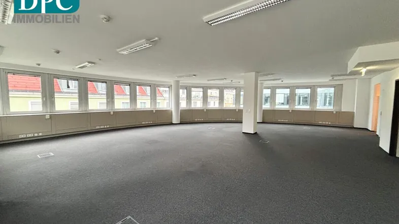 Großer, leerer Büroraum mit Teppichboden und vielen Fenstern, die viel Tageslicht hereinlassen.