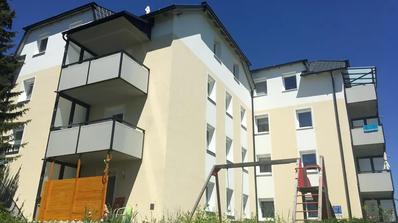 Modernes Mehrfamilienhaus mit hellbeiger Fassade, Balkonen und einem kleinen Spielplatz im Vordergrund.