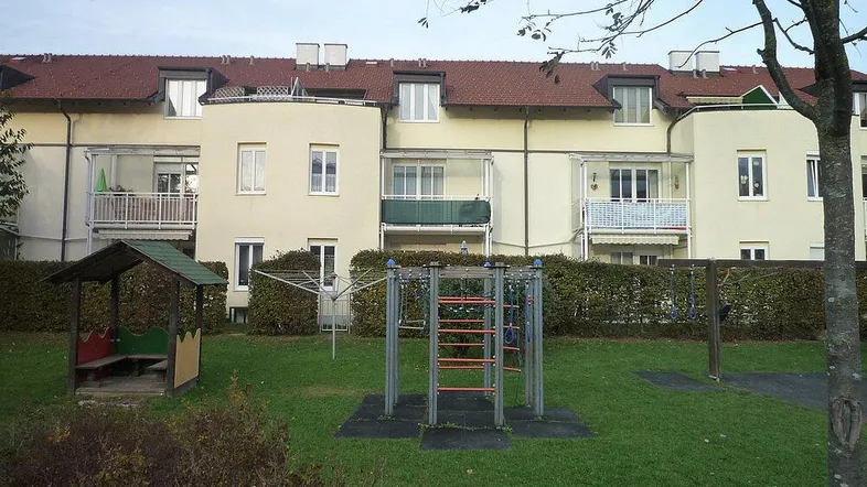 Mehrfamilienhaus mit Balkonen und einem Spielplatz im Vordergrund, umgeben von Grünflächen.