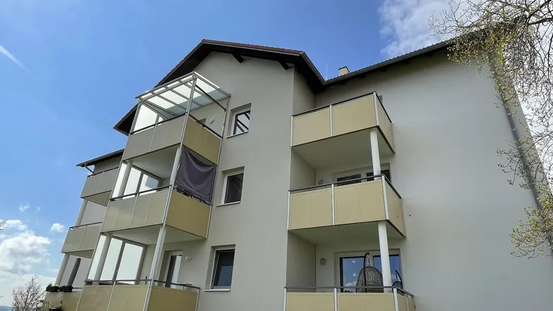 Mehrfamilienhaus mit mehreren Balkonen unter blauem Himmel und grüner Wiese.