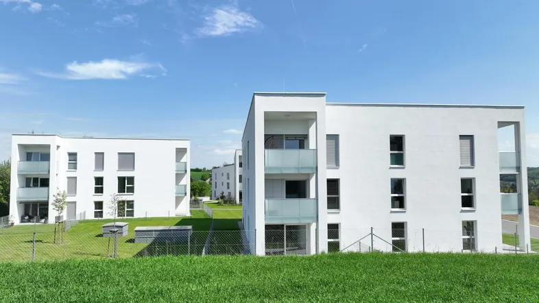 Moderne weiße Mehrfamilienhäuser mit großen Fenstern und grüner Umgebung unter blauem Himmel.