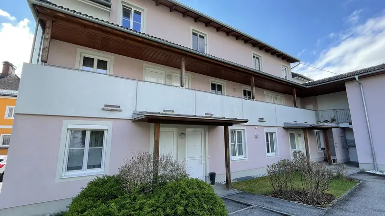 Mehrfamilienhaus mit hellrosa Fassade, Holzbalkonen und gepflegter Grünfläche im Eingangsbereich.