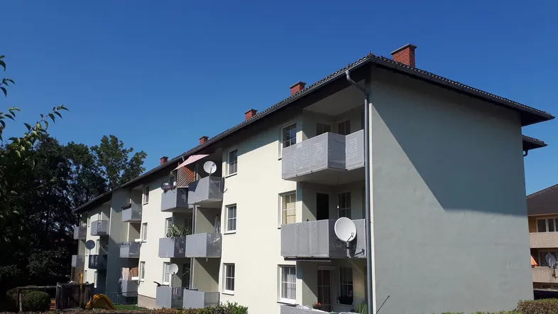 Mehrfamilienhaus mit mehreren Balkonen unter blauem Himmel und grüner Umgebung.