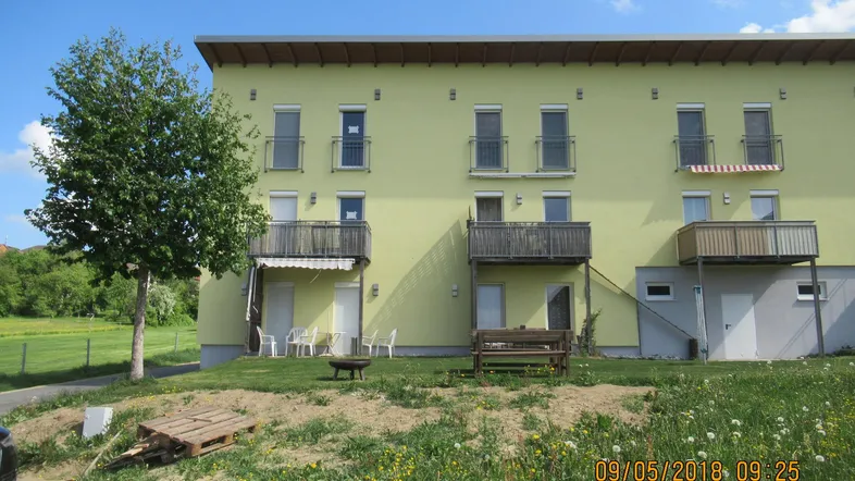 Gelbes Mehrfamilienhaus mit mehreren Balkonen und einer Terrasse, umgeben von einer grünen Wiese.