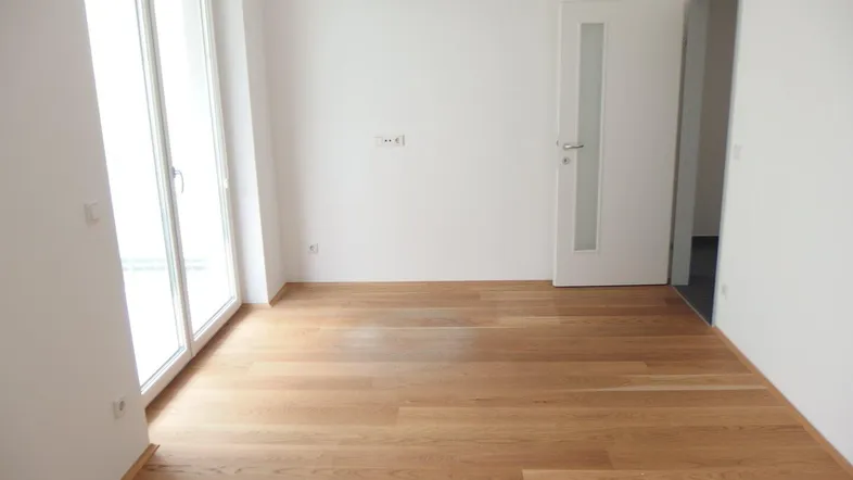 Helles, leeres Zimmer mit Holzboden und Zugang zu einem Balkon oder einer Terrasse.