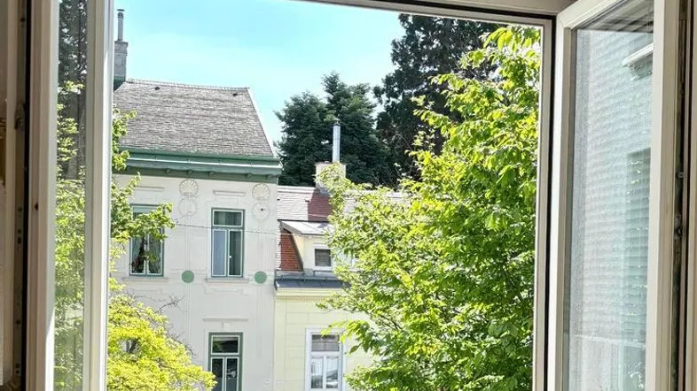 Blick aus einem geöffneten Fenster auf grüne Bäume und umliegende Gebäude.