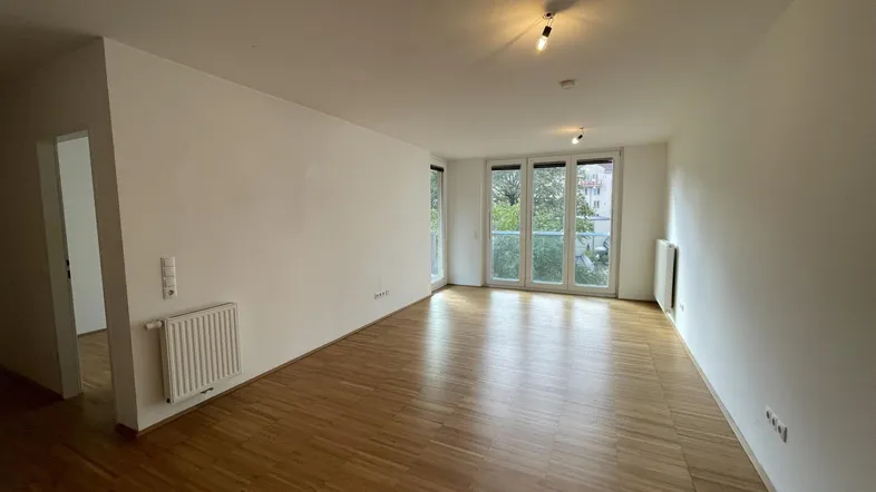 Helles, leeres Wohnzimmer mit Parkettboden und Zugang zu einem Balkon.