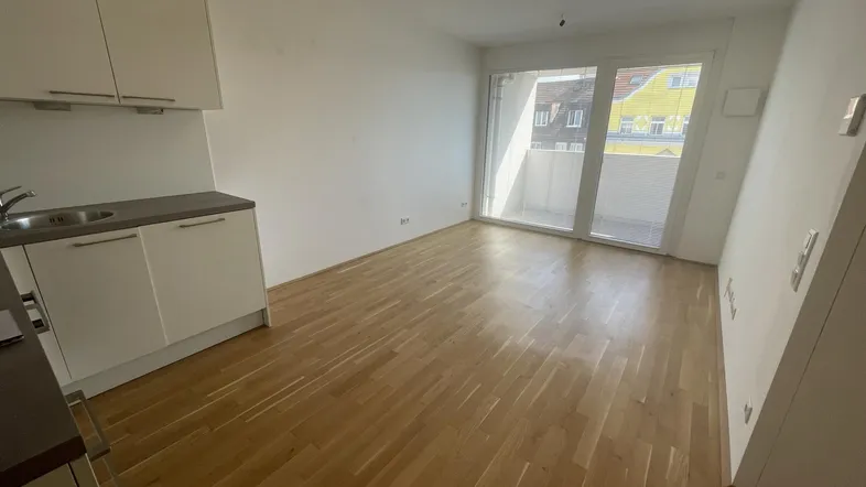Heller Wohnbereich mit integrierter Küchenzeile, Holzboden und großem Balkonzugang.