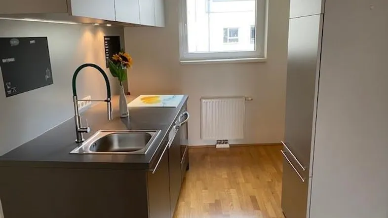 Moderne Küche mit dunklen Unterschränken, hellem Fenster und Holzboden, gut beleuchtet.
