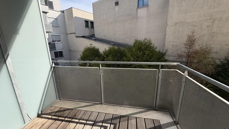 Kleiner Balkon mit Holzboden und Metallgeländer, umgeben von Gebäuden und Bäumen.