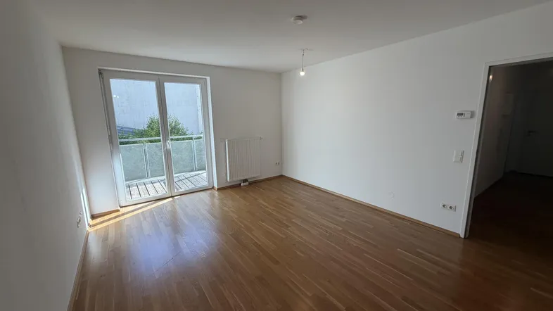 Helles, leeres Wohnzimmer mit Parkettboden und Zugang zu einem kleinen Balkon.