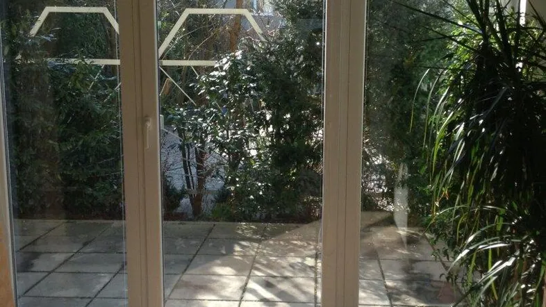 Helles Zimmer mit großen Fenstern und Zugang zu einer gepflasterten Terrasse und Gartenbereich.