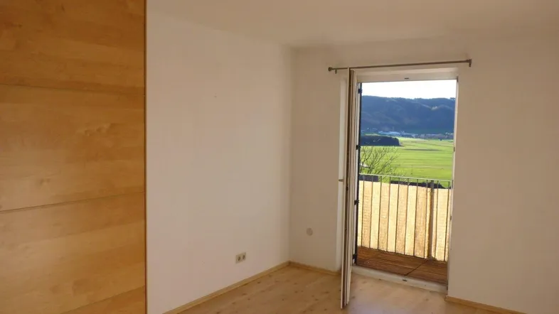 Helles Zimmer mit Holzboden, Einbauschrank und Zugang zu einem Balkon mit weitem Blick ins Grüne.