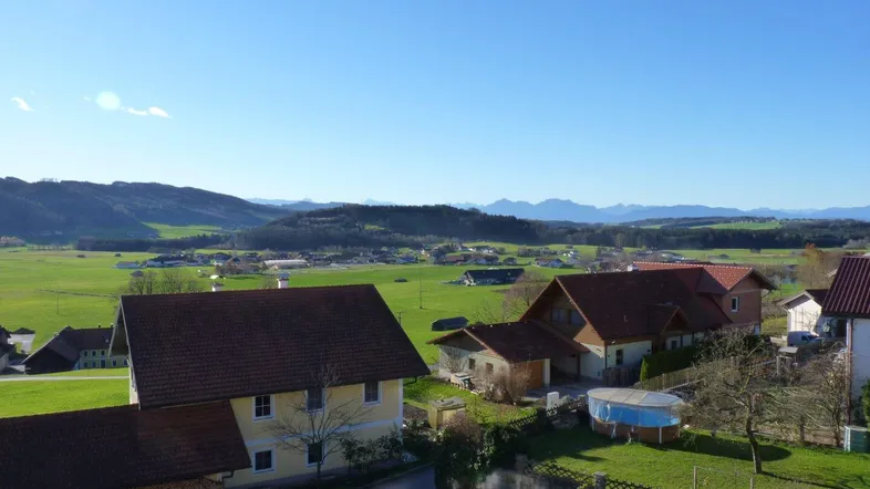 Weitläufige Landschaftsaufnahme mit mehreren Häusern, grünen Feldern und einer Bergkulisse im Hintergrund.