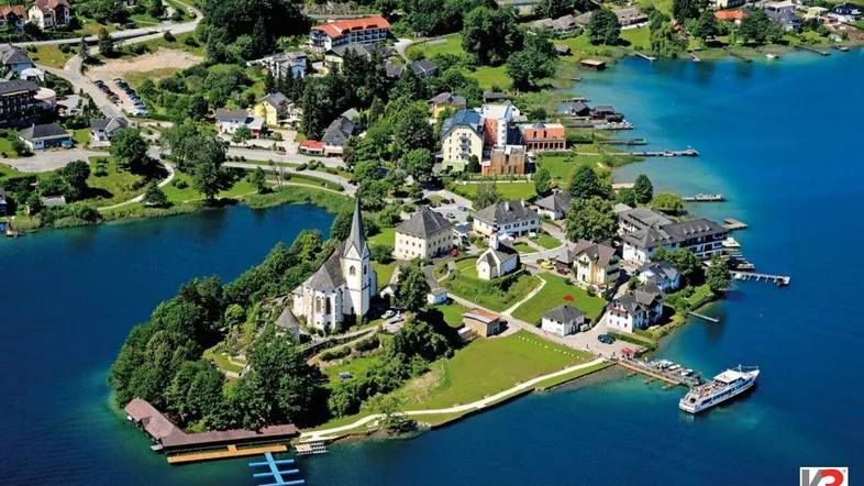 Luftaufnahme eines malerischen Dorfes auf einer Halbinsel am See mit umliegender grüner Landschaft und Bergen.