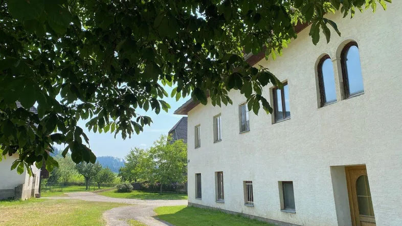 Weiß verputztes Gebäude mit Rundbogenfenstern und Zufahrt, umgeben von grüner Natur und Bäumen.