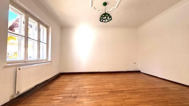 Helles, leeres Zimmer mit Parkettboden, Stuckdecke und großem Fenster.